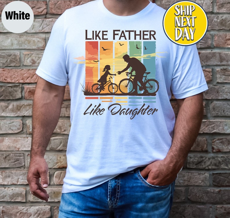 May include: White t-shirt with a retro-style graphic of a father and daughter riding bicycles. The graphic features a sunset background with colorful stripes and the text "Like Father Like Daughter".
