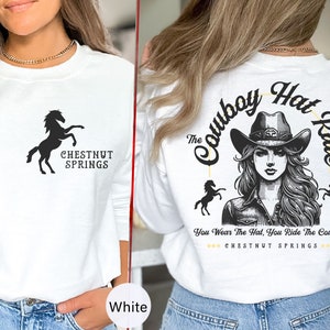 May include: White long-sleeve sweatshirt with a black silhouette of a horse and the text "CHESTNUT SPRINGS". The back of the sweatshirt features a black and white illustration of a woman wearing a cowboy hat with the text "The Cowboy Hat Rule" and "You Wear The Hat, You Ride The Cowboy" with the text "CHESTNUT SPRINGS" underneath.