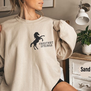 May include: A beige sweatshirt with a black silhouette of a horse and the text "CHESTNUT SPRINGS". The sweatshirt is being worn by a person. The background is a bedroom with a nightstand and a lamp.