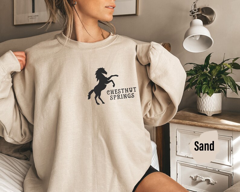 May include: A beige sweatshirt with a black silhouette of a horse and the text "CHESTNUT SPRINGS". The sweatshirt is being worn by a person. The background is a bedroom with a nightstand and a lamp.