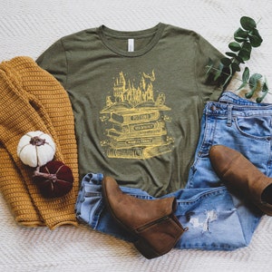 May include: A green t-shirt featuring a yellow graphic of a stack of books with the words "Potions", "Transfiguration", "Charms", and "Herbology" on the spines. The graphic also includes a castle and other magical elements.