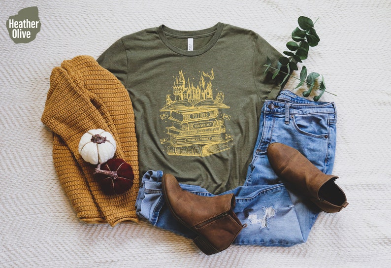 May include: A green t-shirt featuring a yellow graphic of a stack of books with the words "Potions", "Transfiguration", "Charms", and "Herbology" on the spines. The graphic also includes a castle and other magical elements.