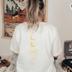 May include: White t-shirt with a graphic of a crescent moon and stars on the back. The moon is repeated in a vertical line with a floral design between each moon.