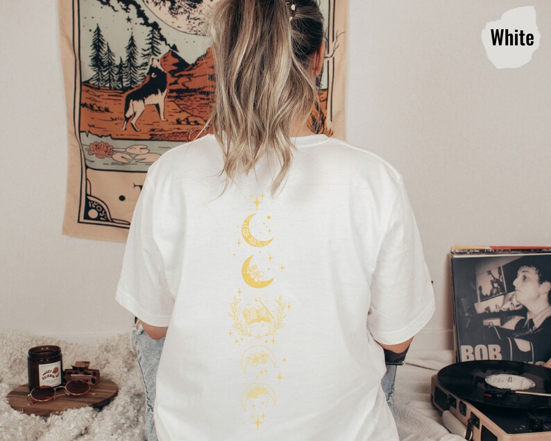 May include: White t-shirt with a graphic of a crescent moon and stars on the back. The moon is repeated in a vertical line with a floral design between each moon.