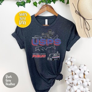 May include: Dark grey heather t-shirt with a graphic that reads "Protect the USPS" in red and blue. The shirt also features a cartoon image of a mail carrier and a mail truck. The text "Keep Public Services Public!" is also included.