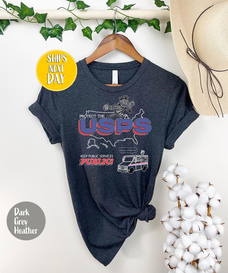 May include: Dark grey heather t-shirt with a graphic that reads "Protect the USPS" in red and blue. The shirt also features a cartoon image of a mail carrier and a mail truck. The text "Keep Public Services Public!" is also included.