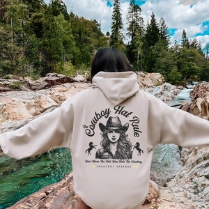 May include: A beige hoodie with a graphic print of a woman wearing a cowboy hat and the text "The Cowboy Hat Rule" and "You Wear The Hat, You Ride The Cowboy" and "CHESTNUT SPRINGS".