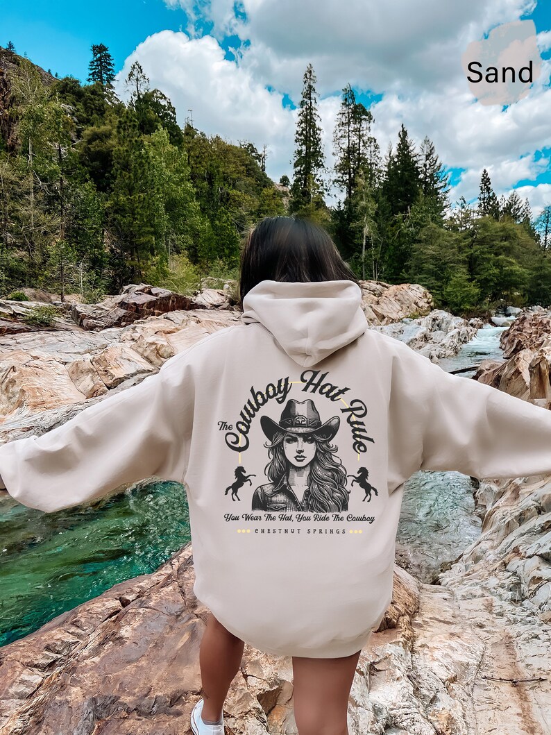 May include: A beige hoodie with a graphic print of a woman wearing a cowboy hat and the text "The Cowboy Hat Rule" and "You Wear The Hat, You Ride The Cowboy" and "CHESTNUT SPRINGS".