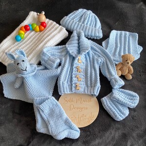 May include: A blue knitted baby set including a cardigan, hat, booties, and a knitted toy. The cardigan has wooden buttons shaped like giraffes. The set is laid out on a dark surface with a wooden sign that says "Silk Marsh Designs".