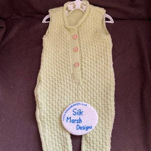 Pode incluir: Um macacão de bebé de malha verde claro com fecho de botões e design sem mangas. O macacão está pendurado num cabide branco. Uma pedra branca com o texto "Silk Marsh Designs" está no canto inferior direito da imagem.