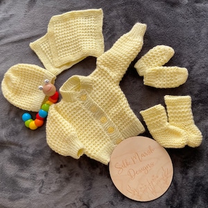 May include: A yellow knitted baby set including a cardigan, hat, booties, and mittens. The set is made with a textured stitch and has a button closure on the cardigan. The set is laid out on a gray surface with a wooden sign that says "Silk Marsh Designs".