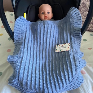 Op de afbeelding: Een babypop zit in een autostoeltje, gewikkeld in een lichtblauwe gebreide deken. De deken heeft een ribbelpatroon en een geschulpte rand. Een label met de tekst "Designed by S. Marsh" is bevestigd.