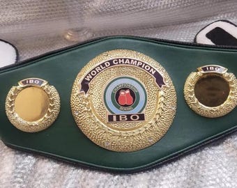 Könnte beinhalten: Ein grüner Lederboxgürtel mit goldenen Akzenten. Der Gürtel trägt die Aufschrift "World Champion" und "IBO". Der Gürtel hat drei goldene Medaillons mit den Buchstaben "IBO" darauf.