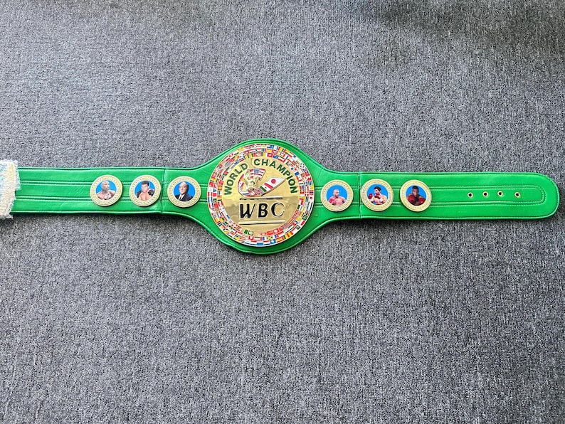 K&ouml;nnte beinhalten: Ein gr&uuml;ner und goldener Box-Championschaftsg&uuml;rtel mit den Worten "World Champion" und "WBC" auf der Vorderseite. Der G&uuml;rtel hat mehrere kleine Flaggen um den Mittelkreis herum und zeigt Portr&auml;ts von Boxern.