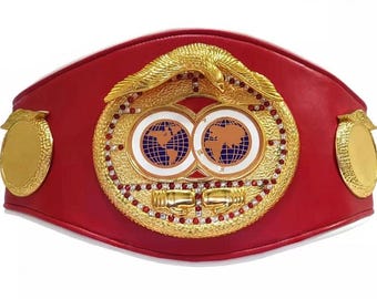 Peut inclure: Une ceinture de champion de boxe rouge et or avec une grande plaque centrale ornée représentant deux globes et un aigle doré. La ceinture a deux petites plaques dorées sur les côtés.
