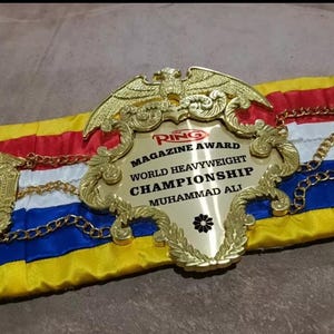 May include: A gold boxing championship belt with a red, white and blue ribbon. The belt features a large gold eagle and the text "Ring Magazine Award World Heavyweight Championship Muhammad Ali".
