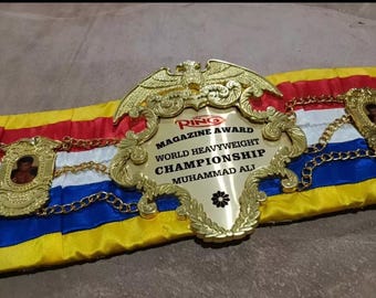 Könnte beinhalten: Ein goldener Box-Championschaftsgürtel mit einem rot-weiß-blauen Band. Der Gürtel zeigt einen großen goldenen Adler und den Text "Ring Magazine Award World Heavyweight Championship Muhammad Ali".