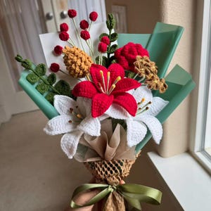 Può includere: Un bouquet all'uncinetto fatto a mano con gigli rossi e bianchi, bacche rosse e altri elementi strutturati. Il bouquet è avvolto in carta verde e marrone, legato con un nastro verde. Un'arrangiamento floreale unico e duraturo.