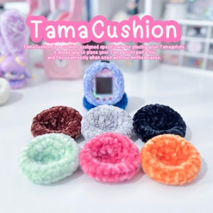 May include: A set of seven colourful crocheted cushions in various colours, including brown, grey, black, green, pink, and orange. The cushions are designed to hold a Tamagotchi, a virtual pet device. The text "Tama Cushion" is written in pink letters on a white background.