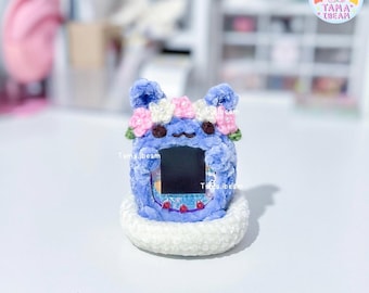 May include: A blue crocheted Tamagotchi toy with a white and pink flower crown. The toy has a black screen and is sitting on a white crocheted base.