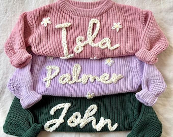May include: Three knitted sweaters with embroidered names, "Isla", "Palmer", and "John", in pink, purple, and green.