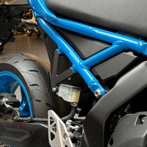 May include: Close-up of a blue motorcycle frame with a black seat and a clear brake fluid reservoir. The motorcycle has a black rear tire with a blue rim.