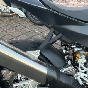 Può includere: Primo piano di una moto nera con un tubo di scarico argentato e uno scudo termico in fibra di carbonio. Sono visibili il pneumatico posteriore e i componenti delle sospensioni, oltre a una sella e un parafango neri. Un'altra moto è parzialmente visibile sullo sfondo.