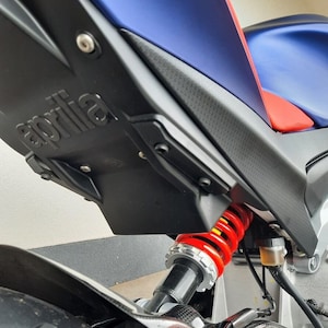 Puede incluir: Primer plano de la suspensión trasera de una motocicleta azul y roja. La motocicleta tiene un guardabarros trasero de fibra de carbono negro con la palabra "aprilia" en relieve. El amortiguador trasero es rojo con un muelle amarillo.