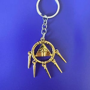 May include: Gold-colored metal keychain featuring the Eye of Anubis symbol with a circle and six spikes. The keychain is attached to a silver metal ring.