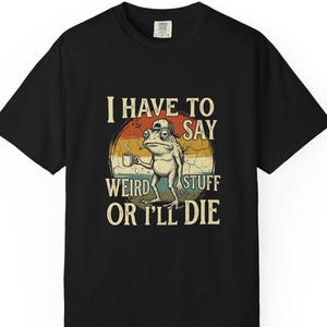 May include: Black t-shirt featuring a cartoon frog with a coffee cup, wearing a hat. The frog is set against a retro sunset design with the text "I HAVE TO SAY WEIRD STUFF OR I'LL DIE."