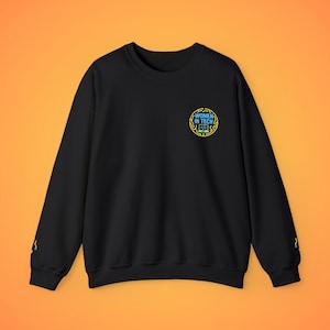 May include: Black crewneck sweatshirt with the text "WOMEN IN TECH" in a blue and yellow circular design on the chest. The sleeves have a small blue and yellow design. The sweatshirt is displayed against an orange background.