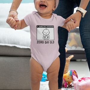 May include: A light pink baby onesie with the text "baby.exe initialized" and "ZERO DAY OLD" printed on the front. The onesie features a graphic of a baby. A baby wearing a pink headband is being held by two adults.