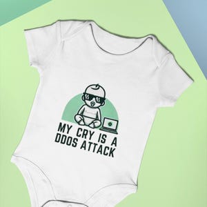 Puede incluir: Un body blanco para bebé con un bebé de dibujos animados verde que lleva gafas de sol y un chupete, sentado frente a un portátil. El texto "MY CRY IS A DDOS ATTACK" está impreso debajo de la imagen.