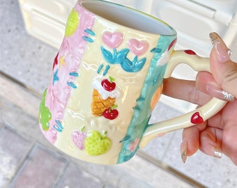 May include: A ceramic mug with a patchwork design featuring hearts, flowers, a cherry ice cream cone, and fruit. The mug is white with pink, blue, and yellow accents.