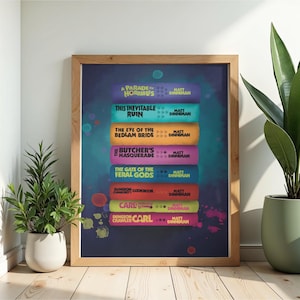 Puede incluir: Un póster enmarcado que muestra una pila de siete lomos de libros coloridos con los títulos A Parade of Horribles, This Inevitable Ruin, The Eye of the Bedlam Bride, The Butcher's Masquerade, The Gate of the Feral Gods, Dungeon Crawler Carl Cookbook y Carl.