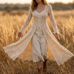 May include: A cream-colored, open-front crochet duster with long sleeves and a lace-up bodice. The duster is worn over a cream-colored dress and pants. The image is taken outdoors in a field of tall grass.