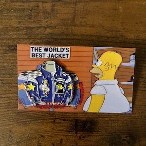 May include: A blue and yellow enamel pin featuring a cartoon jacket with a variety of patches and the text "The World's Best Jacket". The pin is displayed on a card with a cartoon character from the Simpsons.