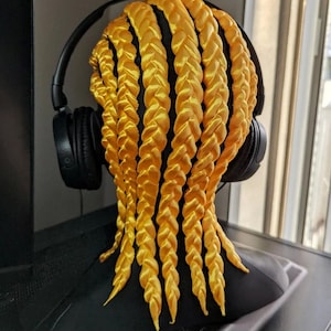 May include: A black mannequin head with long, yellow braided hair. The hair is styled in a series of thick braids that hang down the back of the head. The mannequin is wearing a pair of black headphones.
