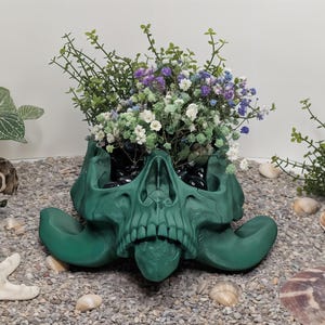 May include: A green skull-shaped planter with a tongue sticking out, filled with white and purple flowers. The planter is sitting on a bed of small gray rocks.