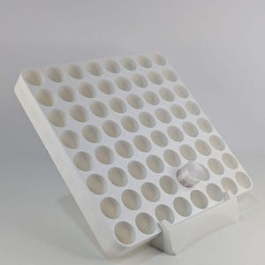May include: A white plastic storage rack with a grid of circular openings arranged in rows and columns, supported by a base stand. One small, clear, cylindrical container with a translucent lid is inserted into one of the circular slots.