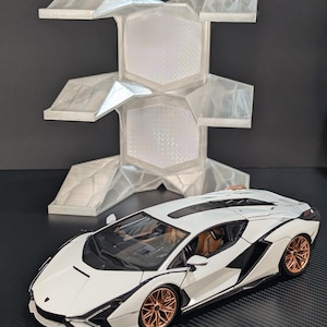 May include: A white Lamborghini Sian model car with copper-colored wheels and a tan interior, positioned in front of a white, multi-tiered, geometric display stand with a textured, crystalline surface finish and hexagonal cutouts.