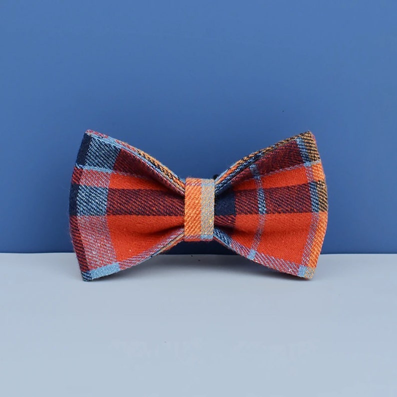 May include: A plaid bow tie in shades of orange, navy, and light blue. The bow tie has a classic shape with a central band in a lighter orange hue. The background is a solid blue.