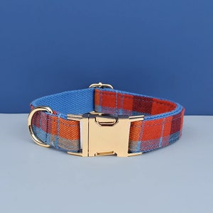 May include: A dog collar with a plaid pattern in shades of orange, red, and blue. The collar has a gold-colored metal buckle and a matching D-ring. The inner lining is a solid blue fabric.