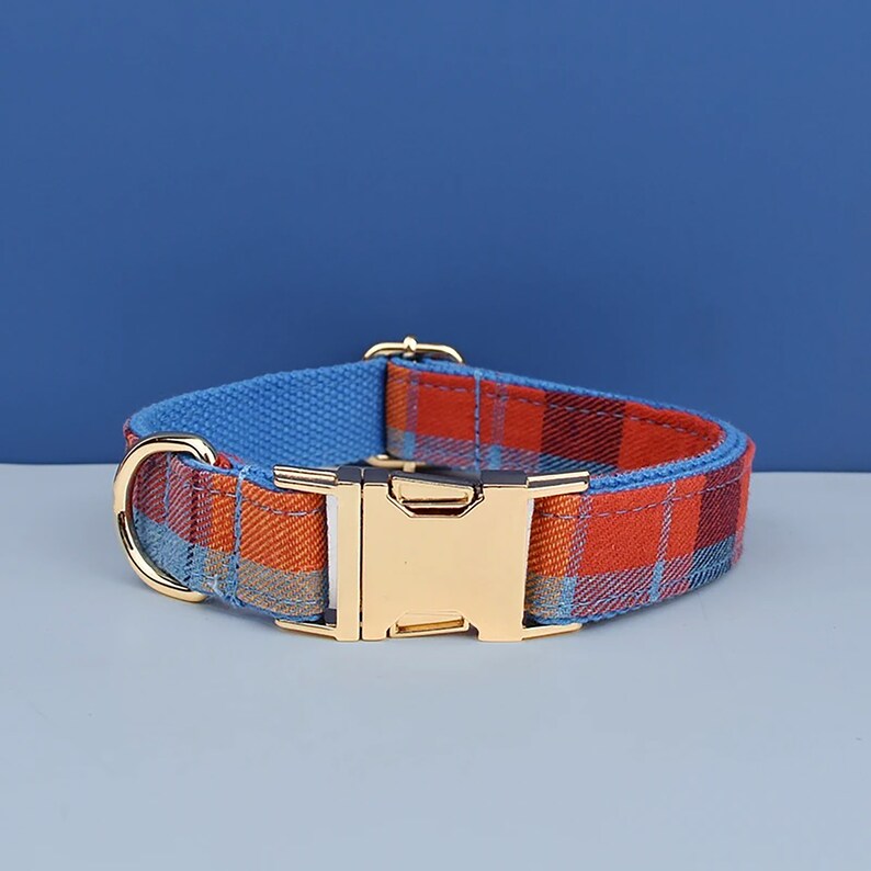 May include: A dog collar with a plaid pattern in shades of orange, red, and blue. The collar has a gold-colored metal buckle and a matching D-ring. The inner lining is a solid blue fabric.