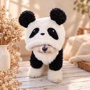 Könnte beinhalten: Ein kleiner Hund trägt einen cremefarbenen Kapuzenpullover aus Plüsch, der wie ein Panda gestaltet ist. Die Kapuze hat schwarze, runde Ohren und schwarze Flecken um die Augen. Der Pullover hat eine weiße Brust und schwarze Ärmel aus weichem, strukturiertem Material.