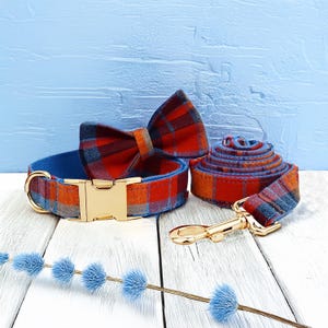 May include: A dog collar, bow tie, and leash set. The collar and leash are a plaid pattern in red, orange, and blue. The collar has a gold-colored metal buckle. The bow tie is a matching plaid. The leash is rolled up.
