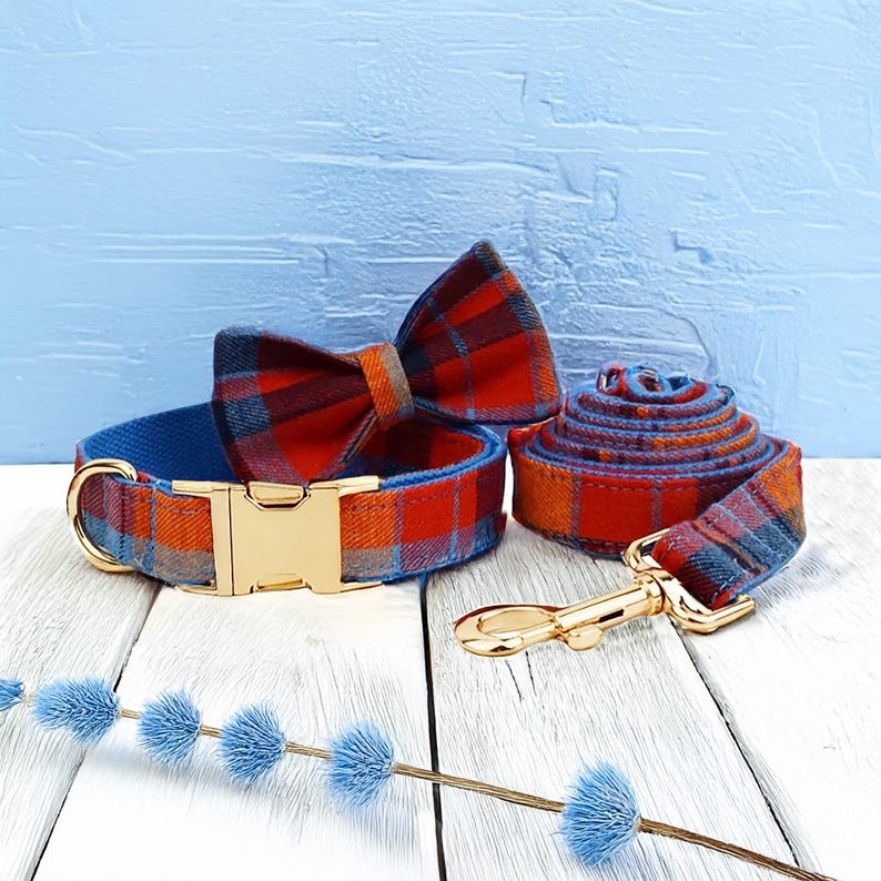 May include: A dog collar, bow tie, and leash set. The collar and leash are a plaid pattern in red, orange, and blue. The collar has a gold-colored metal buckle. The bow tie is a matching plaid. The leash is rolled up.