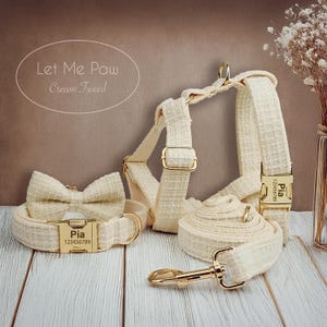 Könnte beinhalten: Cremefarbenes Tweed-Hunde-Set mit Halsband, Geschirr, Leine und Fliege, alle mit goldfarbenen Metallbeschlägen. Halsband und Geschirr haben Namensschilder mit dem Namen "Pia" und einer Telefonnummer. Der Text "Let Me Paw Cream Tweed" ist enthalten.
