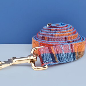 May include: Rolled plaid dog leash in shades of orange, red, and blue with a gold-colored metal clasp. The leash is made of a woven fabric and is designed for pet owners.