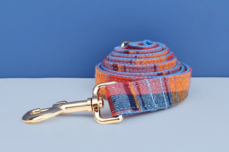 May include: Rolled plaid dog leash in shades of orange, red, and blue with a gold-colored metal clasp. The leash is made of a woven fabric and is designed for pet owners.